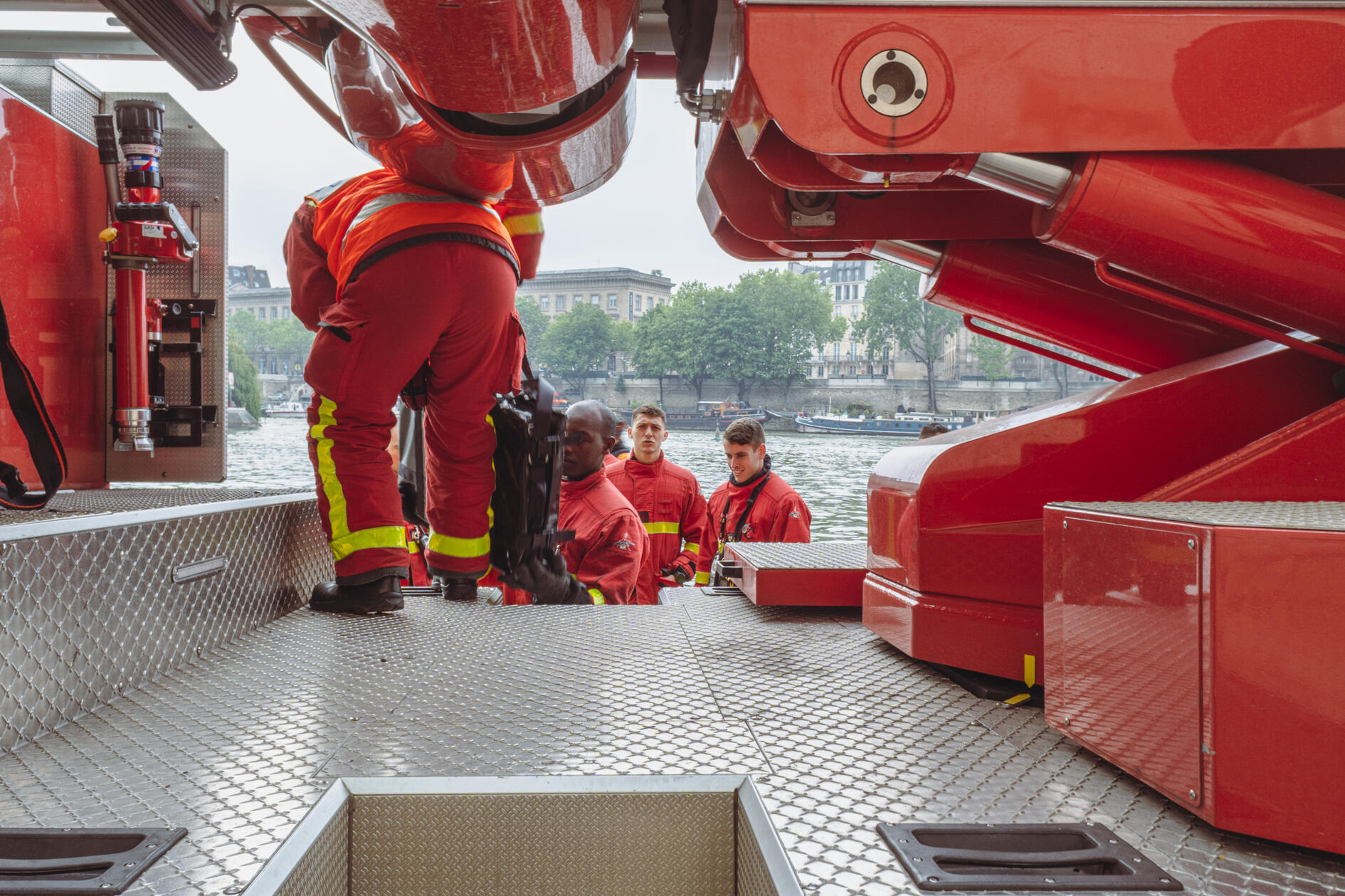 Firefighters stand among the red machinery of a fire truck on a Paris street, one looking directly into the camera.